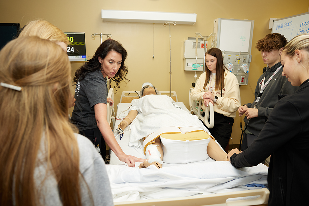 A nursing instructor points to a dummy on a hospital bed in a lab. Students surround her and listen.