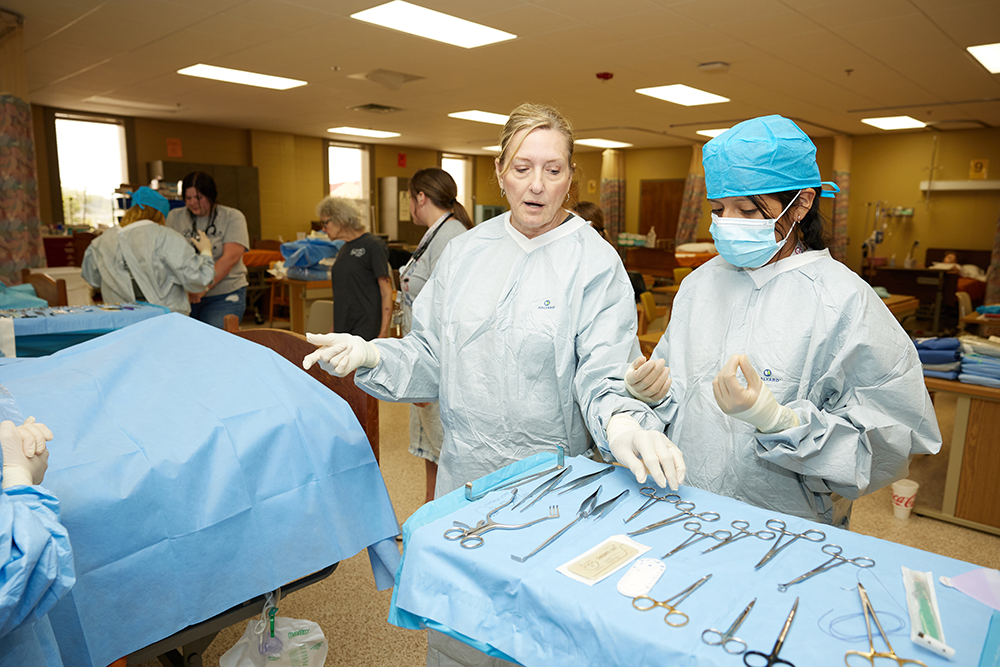 A teacher and student wear scrubs. The teacher points to various medical instruments on a table.