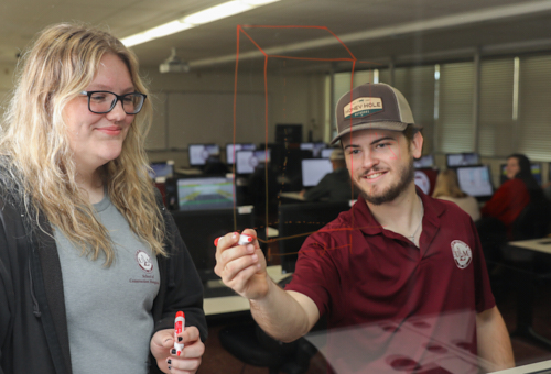 Two ULM Construction Management students draw geometry shapes on a glass in red marker. A computer lab is in the background.