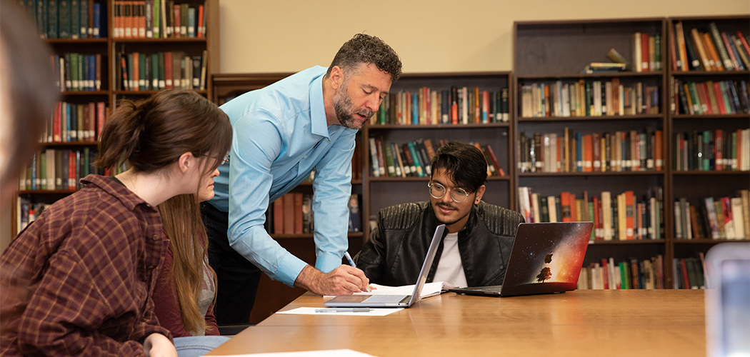 A professor leans on a table and writes on paper. Students look at what he is writing.