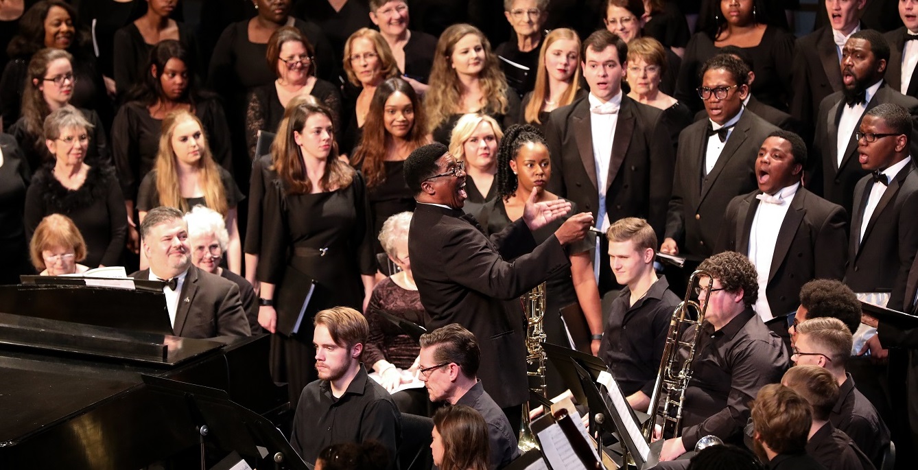 A man conducts a choir and band. They are all wearing black dresses or black suits. They surround him and he is smiling.