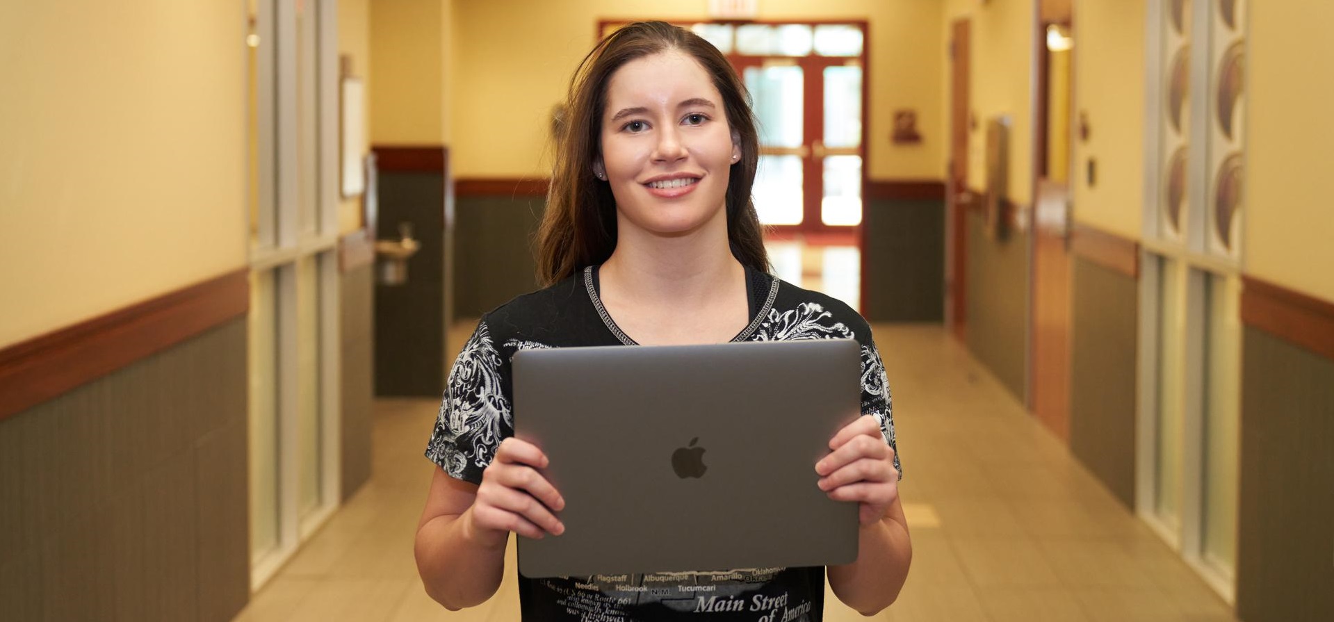 A woman holds a laptop, smiling at the camera, in a hallway.