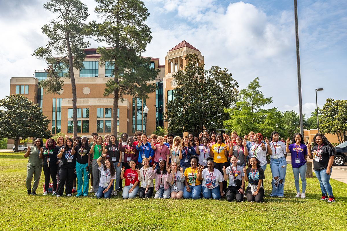 A group of Nursing Bootcamp students stand in a field in front of the ULM Library..
