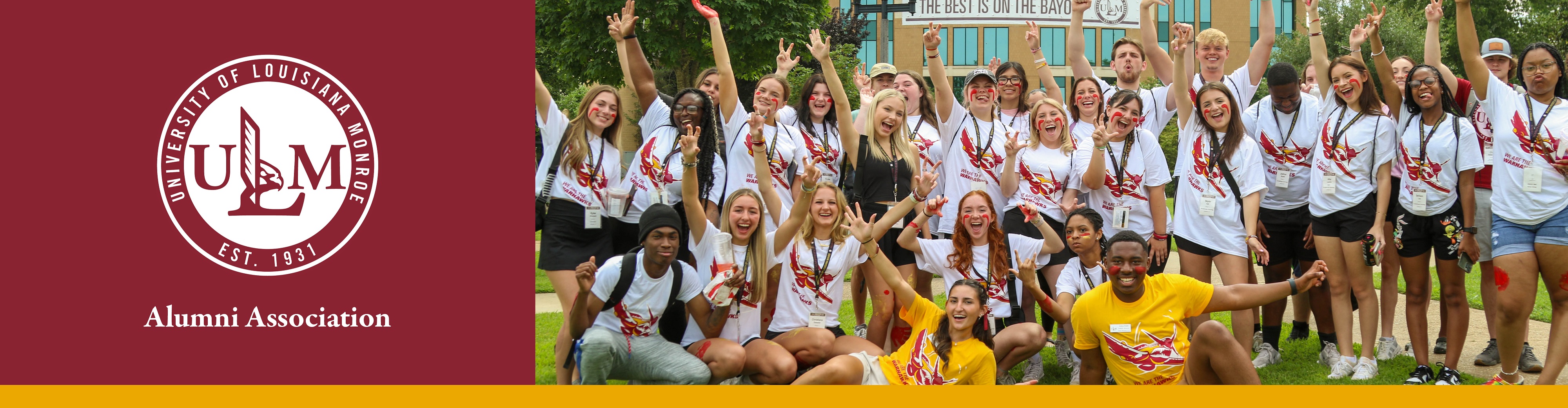 A group of college students cheer and smile at the camera. They are wearing matching shirts and stand in front of a library.