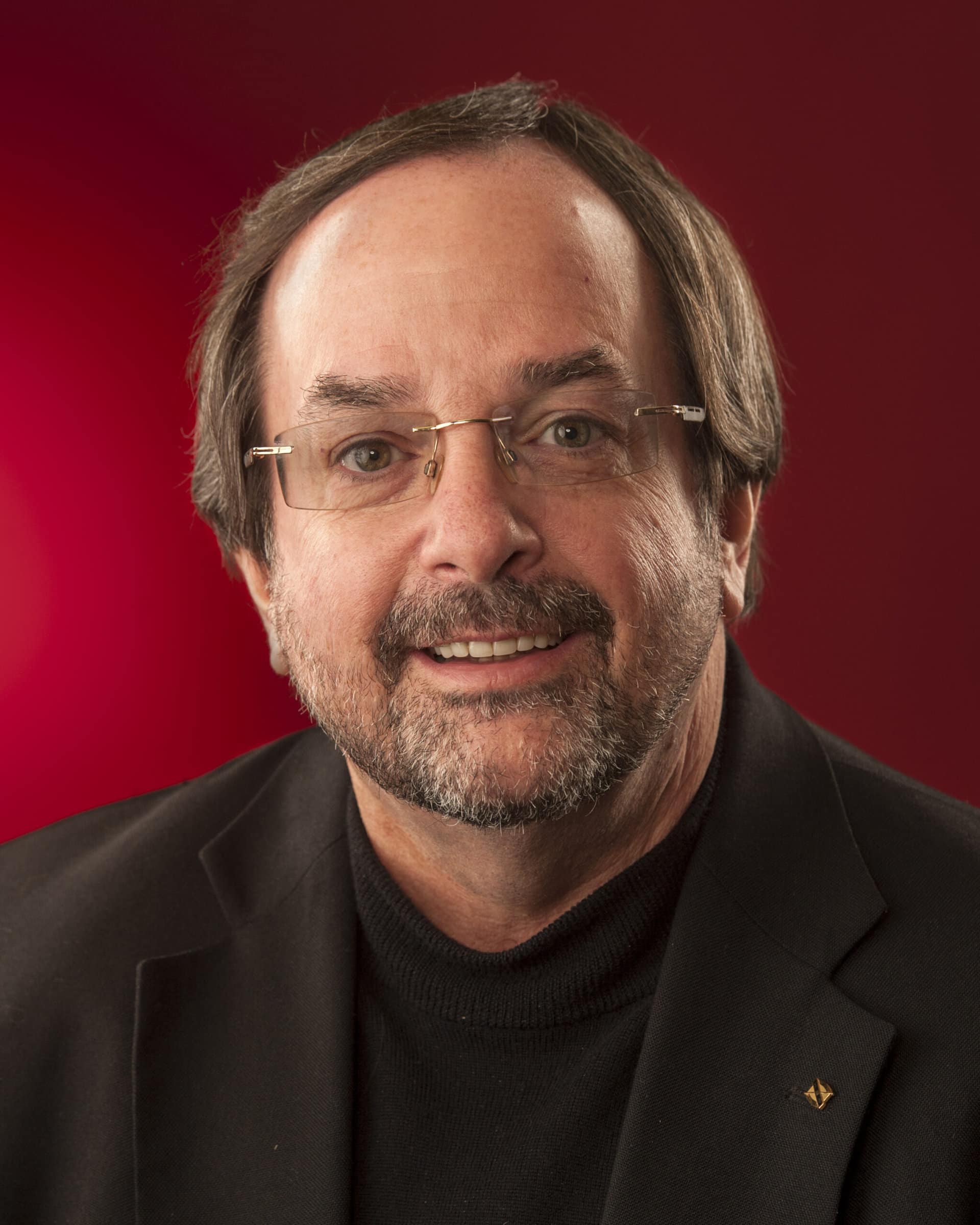 Dr. Larry Blocher is seated in front of a red background and is wearing black clothes.
