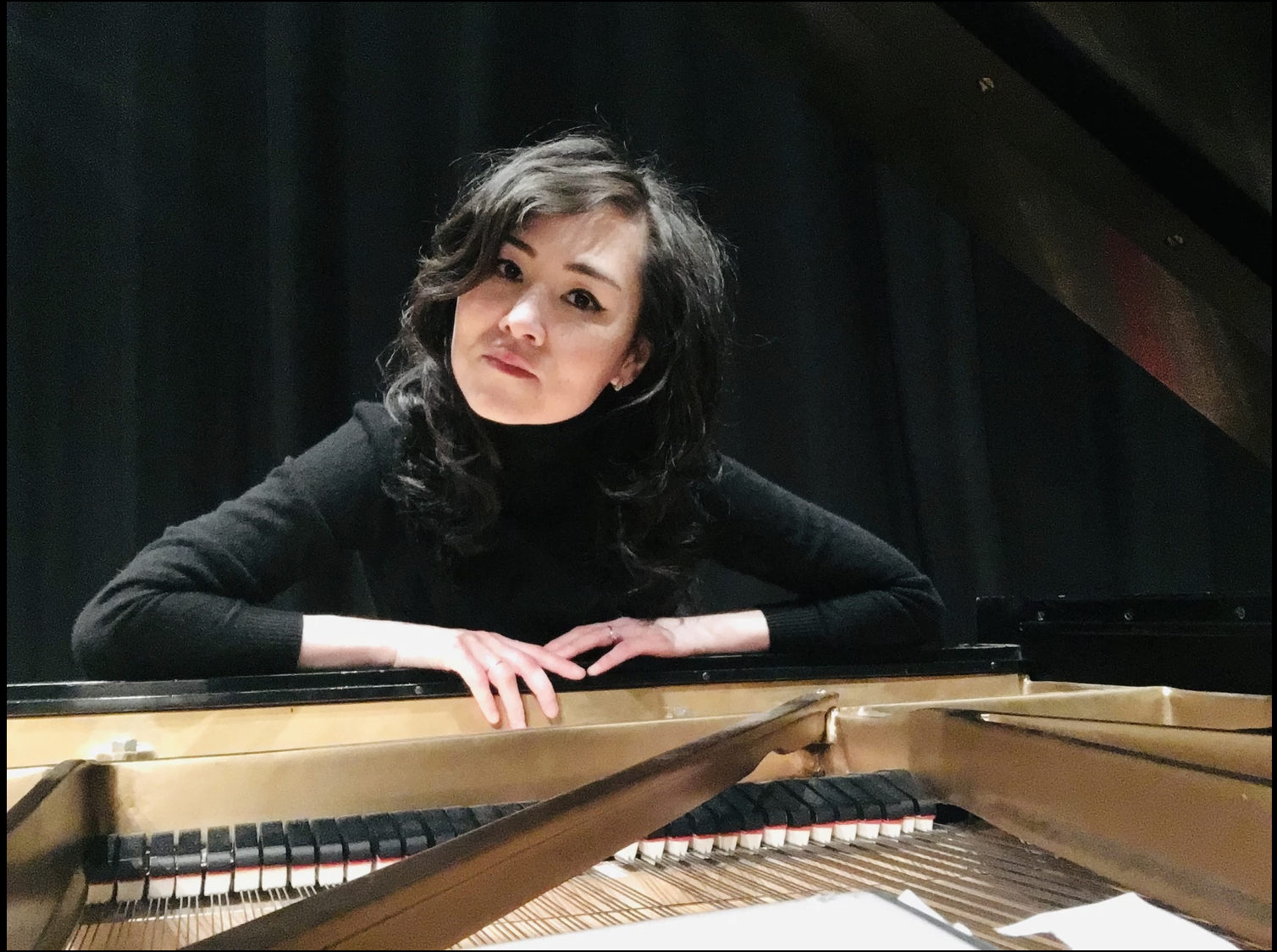 Diana Thacher is seated at a grand piano with the lid open. She is leaning across the keys and the inside of the piano is visible. She is wearing black clothes.
