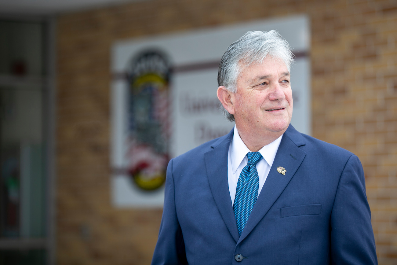 A man stands in front of a brick building. He is smiling and looking to the right of the camera.