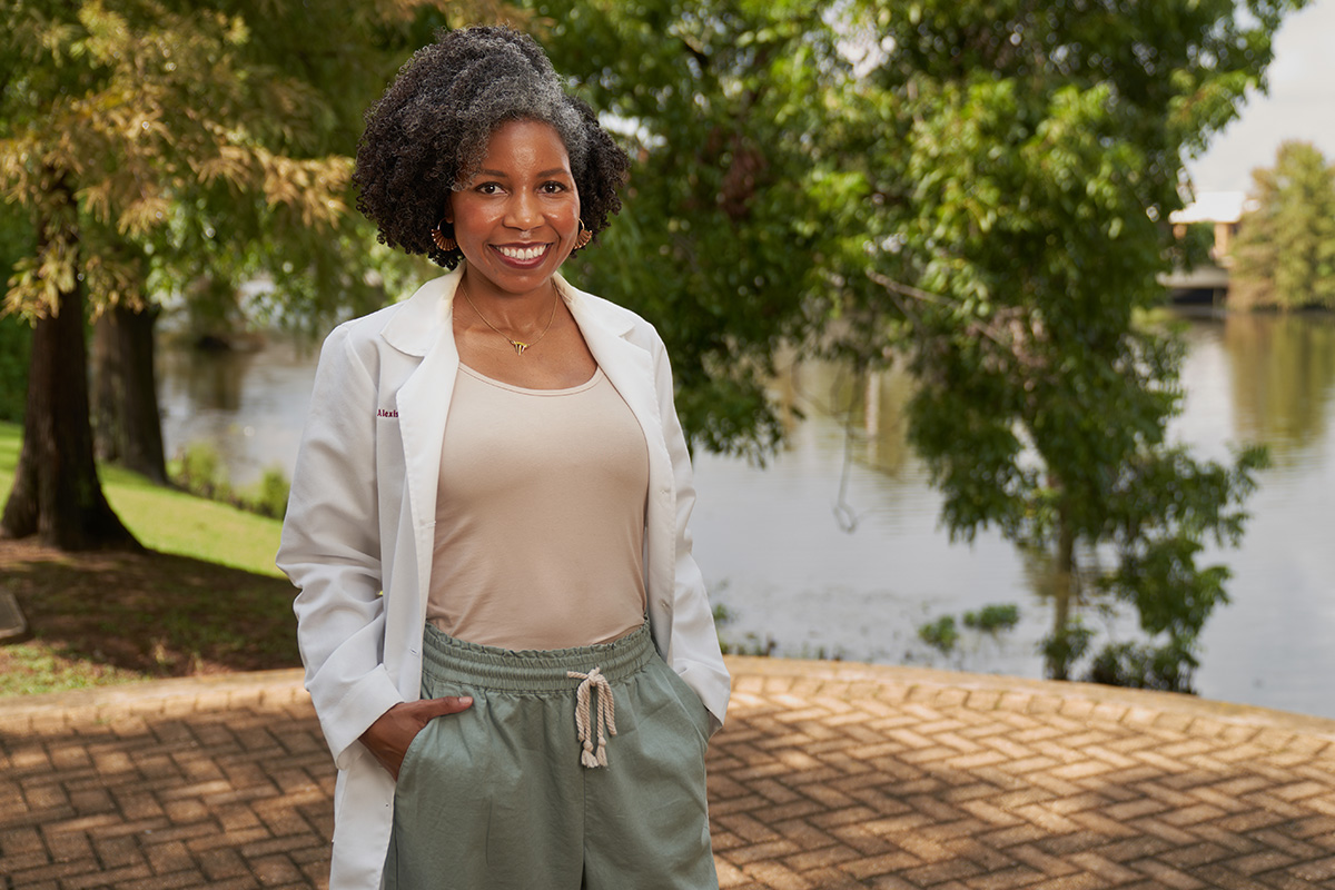 A woman in a white coat is smiling at the camera. She stands in front of a body of water.
