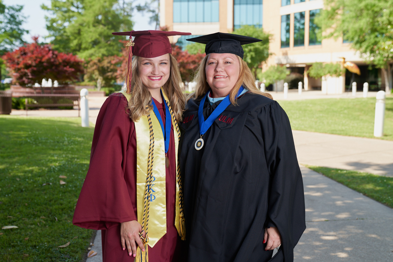 Gabby and Shannon Ballew smile at the camera, wearing their caps and gowns in front of the ULM library.