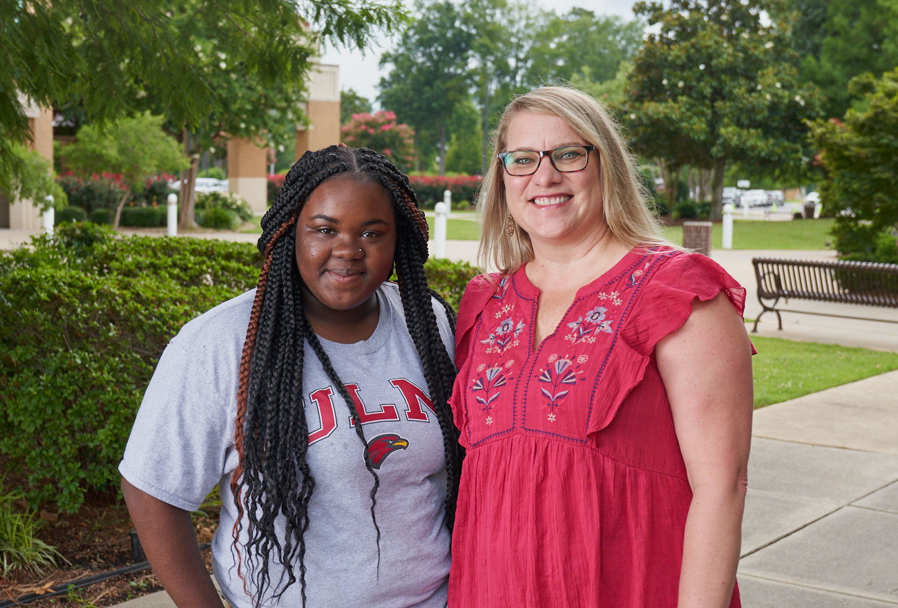 Two women smile at the camera. They are outdoors in front of trees and a plaza.