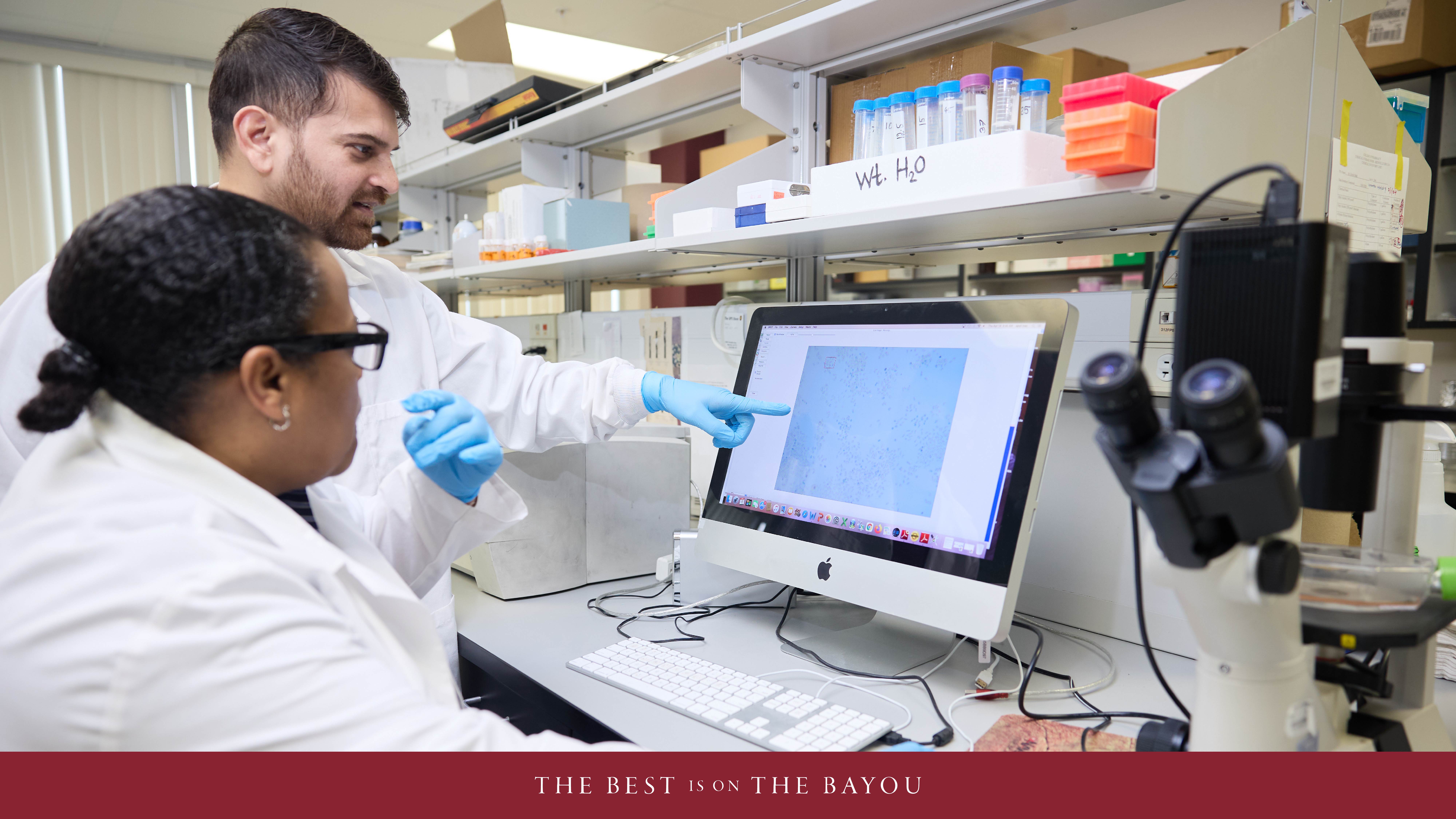 Two students in the ULM College of Pharmacy work in a laboratory setting, analyzing data on a computer screen. 