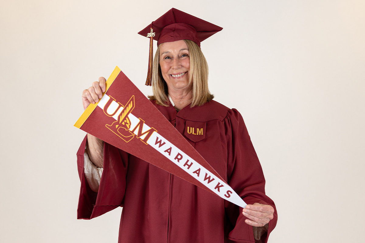 A woman in a ULM graduation cap and gown smiles and holds a ULM Warhawks pennant.