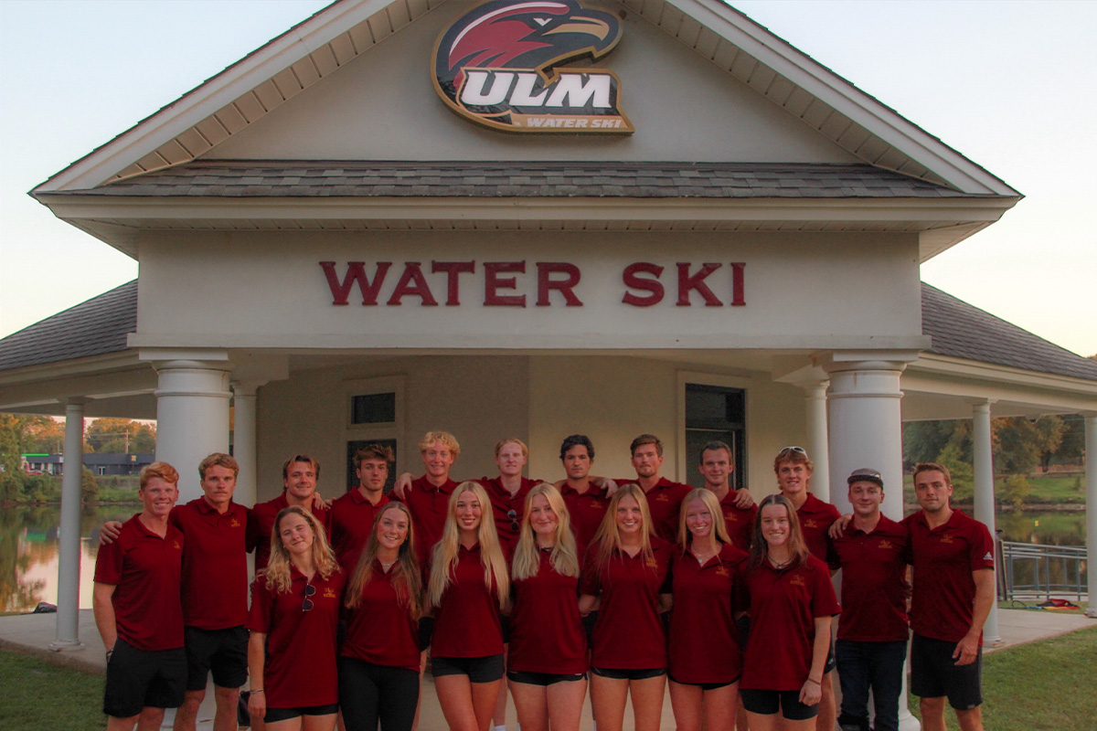 A group of peple linking arms and smiling with the words "ULM Water Ski" on a sign above them.