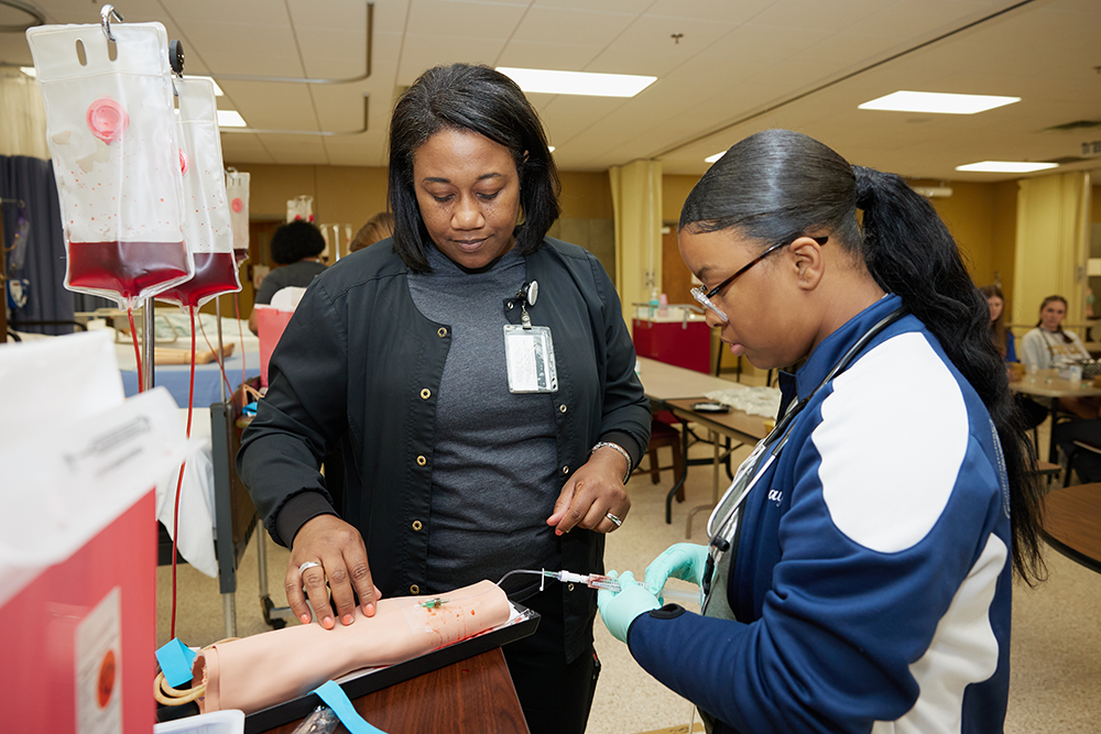 A student draws fake blood from a prosthetic in a classroom lab while her instructor looks on.