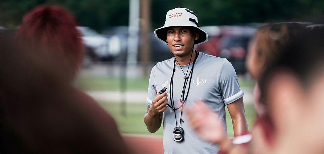 A man in a hat outdoors pulls a whistle to his mouth.